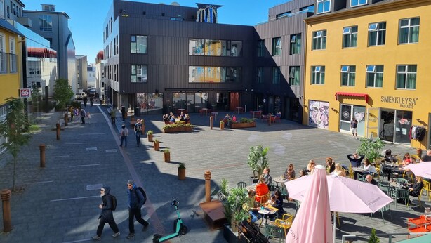 En offentlig plads med moderne bygninger, udendørs siddepladser og både fodgængere og cyklister.