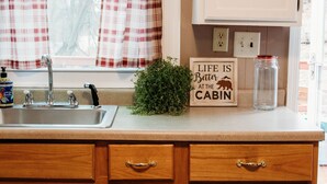Une cuisine équipée d’un évier, de meubles en bois, d’une fenêtre ornée de rideaux à carreaux, d’une plante en pot et d’une pancarte sur laquelle on peut lire : « La vie est plus belle à la cabane ».