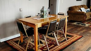 Un coin repas avec une table en bois, quatre chaises, une horloge au mur, une porte à l’arrière-plan et un tapis à motifs sur le sol.
