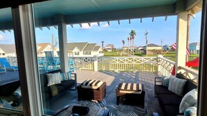A covered outdoor deck with wicker furniture, a white picket fence, and a view of houses and palm trees.