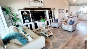 A spacious living room with a large TV, comfortable seating, and a kitchen in the background.