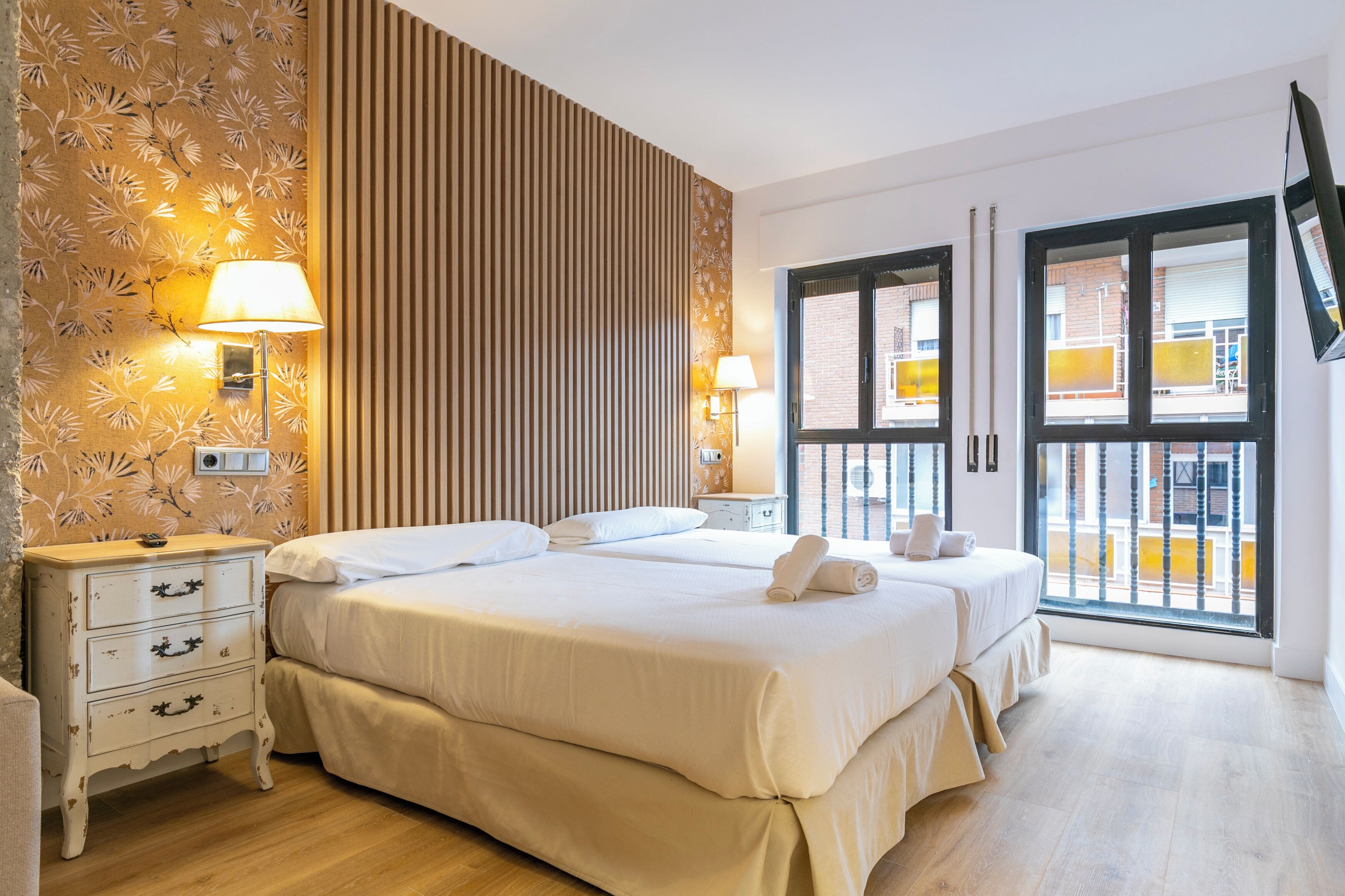 Una camera d'albergo con due letti, un comodino, una lampada, un televisore fissato al muro e una finestra vista esterna.