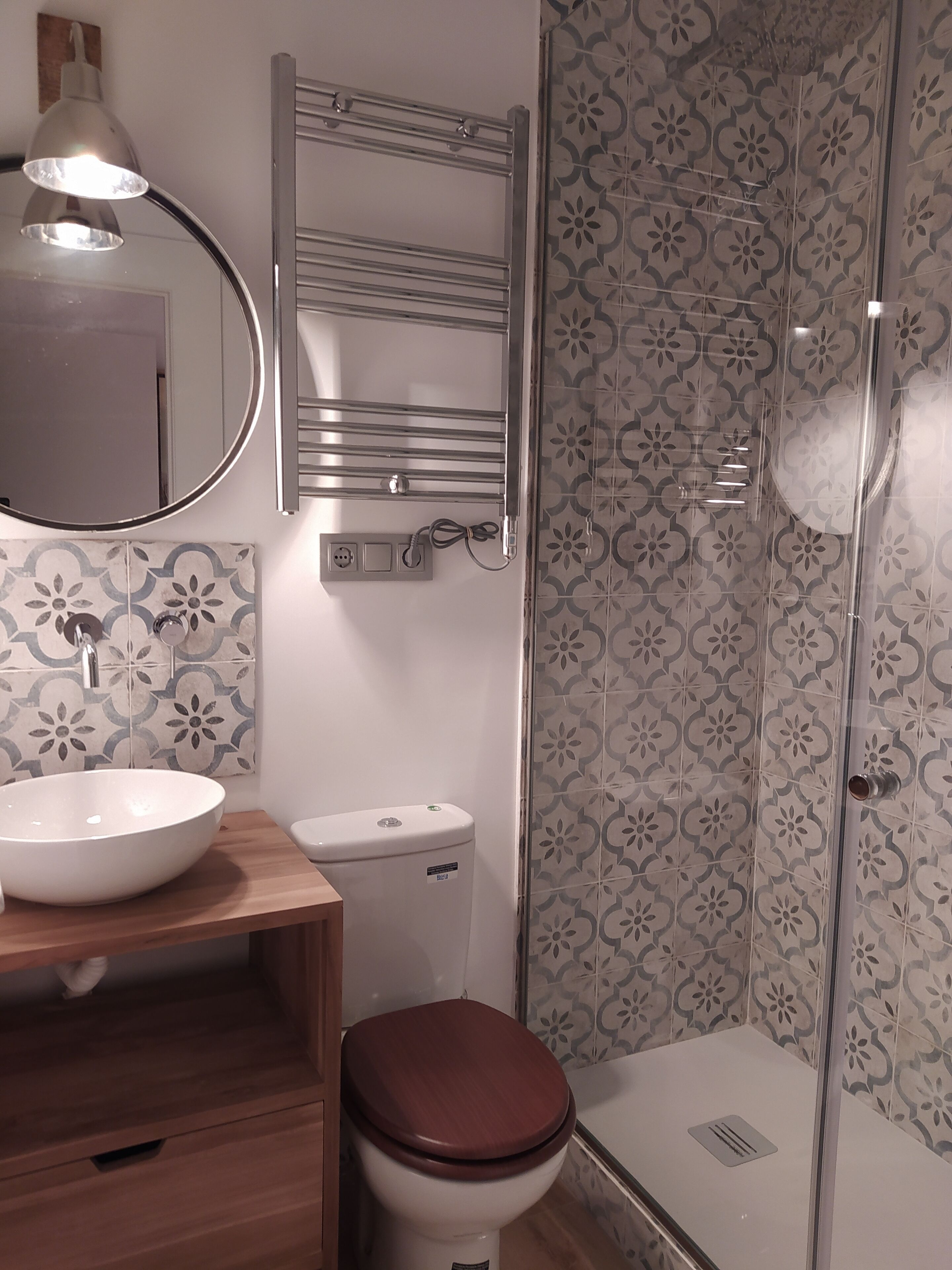 Un baño con tocador de madera, un lavamanos blanco, un inodoro, un espejo y un box de ducha con azulejos estampados.