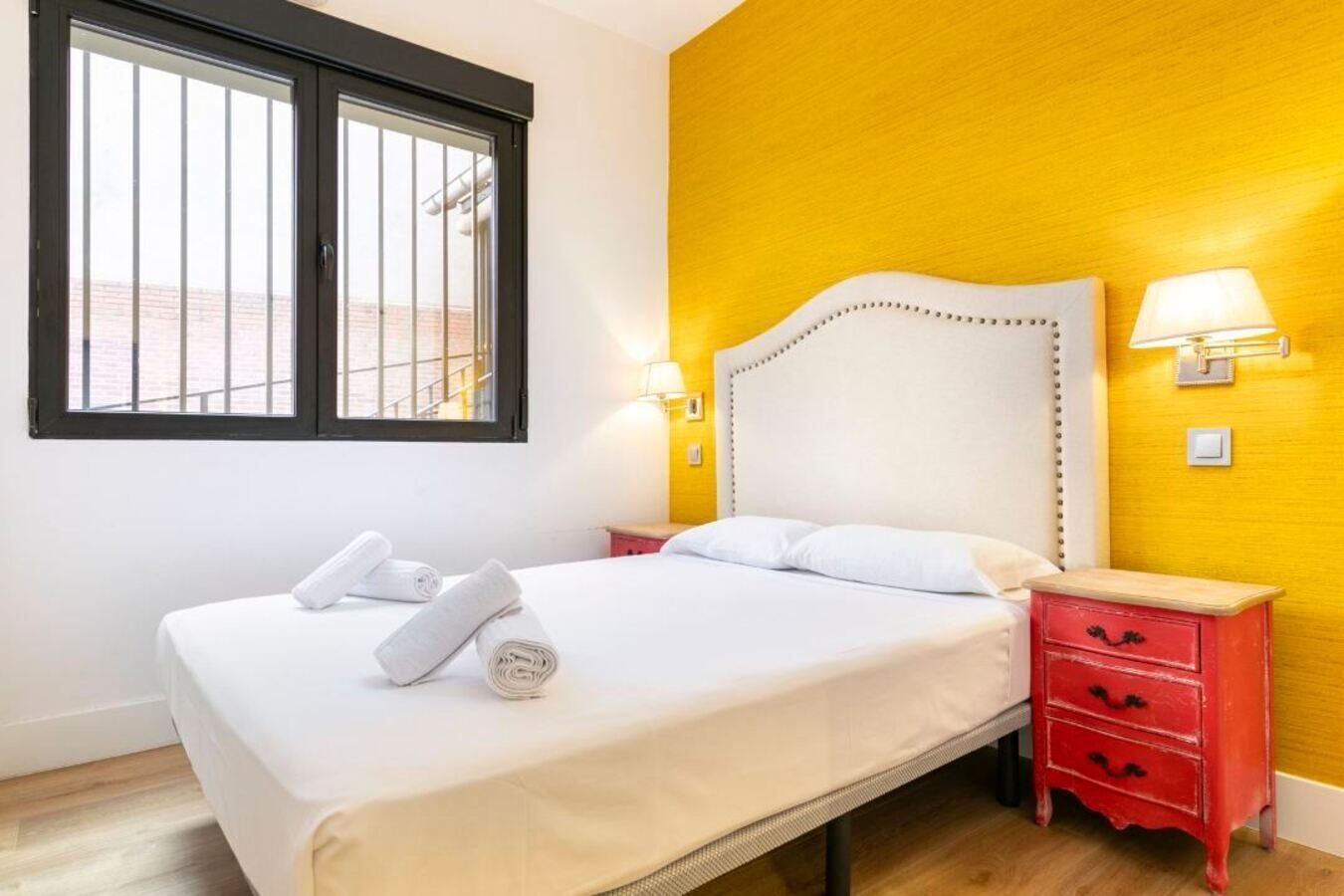 Una habitación de hotel con una cama, dos mesitas de noche, un ventanal y una pared amarilla.