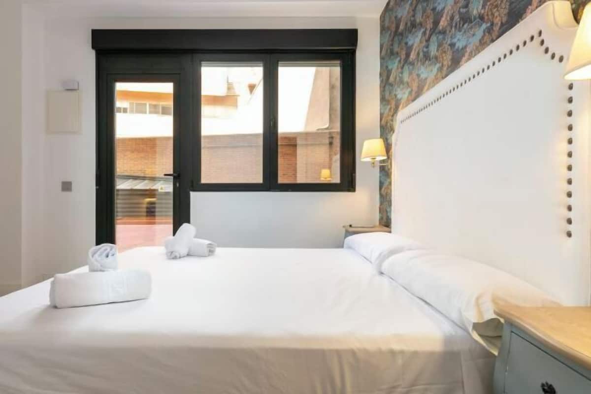 A hotel room with a bed, a nightstand, a lamp, a window, and a door.