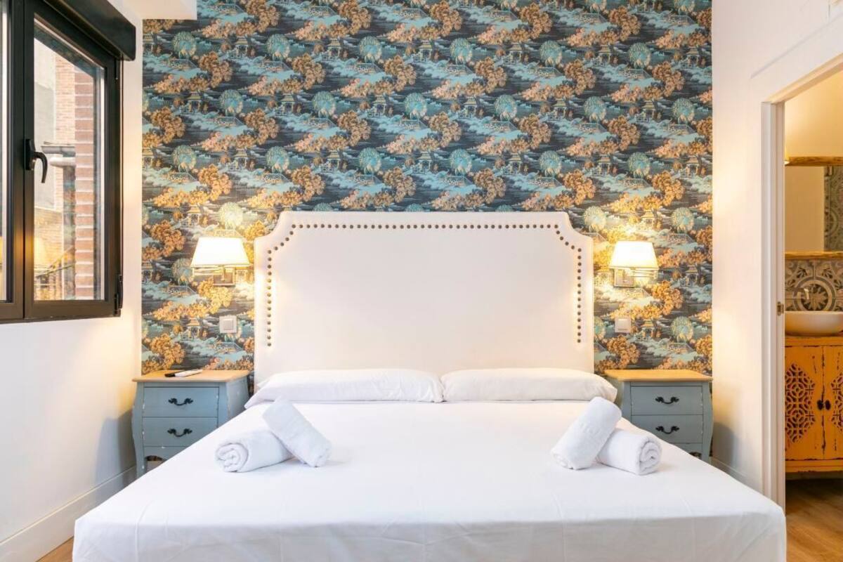 A hotel room with a bed, two bedside tables, a nightstand, a window, and a wall with a floral wallpaper pattern.