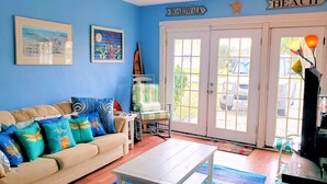 A room with a blue wall, a beige sofa with colorful pillows, a rocking chair, a wooden coffee table, and a door leading outside. - Eel Creek Escape -  Water Front  - Water Access - Screened Porch NO LINENS (Chincoteague)