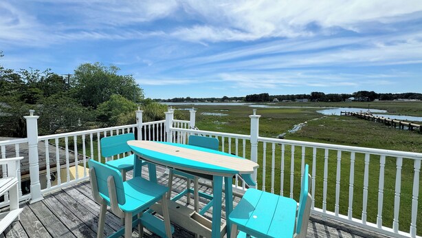 A wooden deck with turquoise chairs and a table, overlooking a grassy area with a water body and distant trees. - Eel Creek Escape -  Water Front  - Water Access - Screened Porch NO LINENS (Chincoteague)