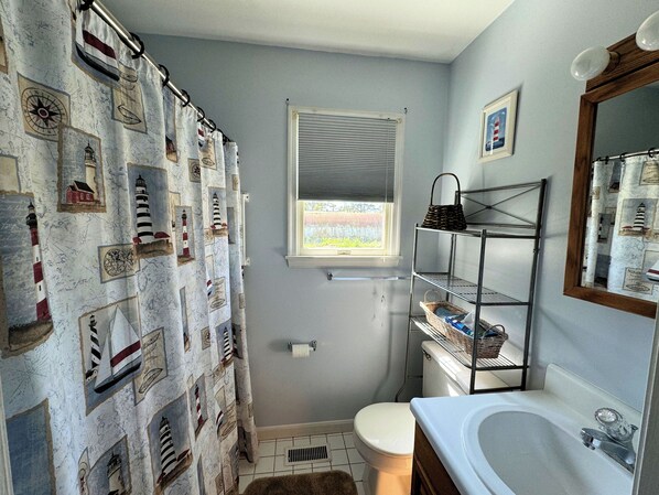 A bathroom with a nautical-themed shower curtain, a window with blinds, a toilet, a shelf with toiletries, and a mirror with a decorative frame.