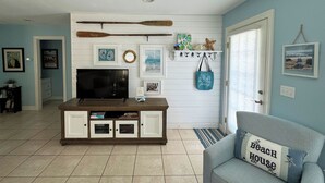 A living room with a TV, books, and a coffee table. There is a blue couch with a pillow that says "Beach House". - Josie's Getaway - Pet Friendly -Mini-Vacations and Month to Monthly Available (Chincoteague)