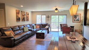 A spacious living room with a leather sectional sofa, a wooden coffee table, and a wooden dining table. There is a ceiling fan, a large window with a view of trees, and artwork on the walls. - A/C LakeAccess GameRoom Xbox DogOK kayaks paddleboards Private30'Dock Sleeps 14 (Sagle)