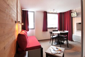 Une pièce de taille modeste avec un mur en bois, un canapé rouge, une petite table avec des magazines, une table à manger avec des chaises et un téléviseur fixé au mur.