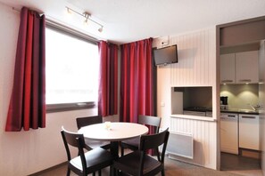 Ein kleines Zimmer mit Esstisch und Stühlen, einem an der Wand befestigten Fernseher und einer Küchenzeile.
