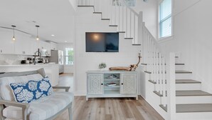Una cocina moderna con armarios blancos, un comedor con un banco de color claro y una escalera con barandillas blancas.