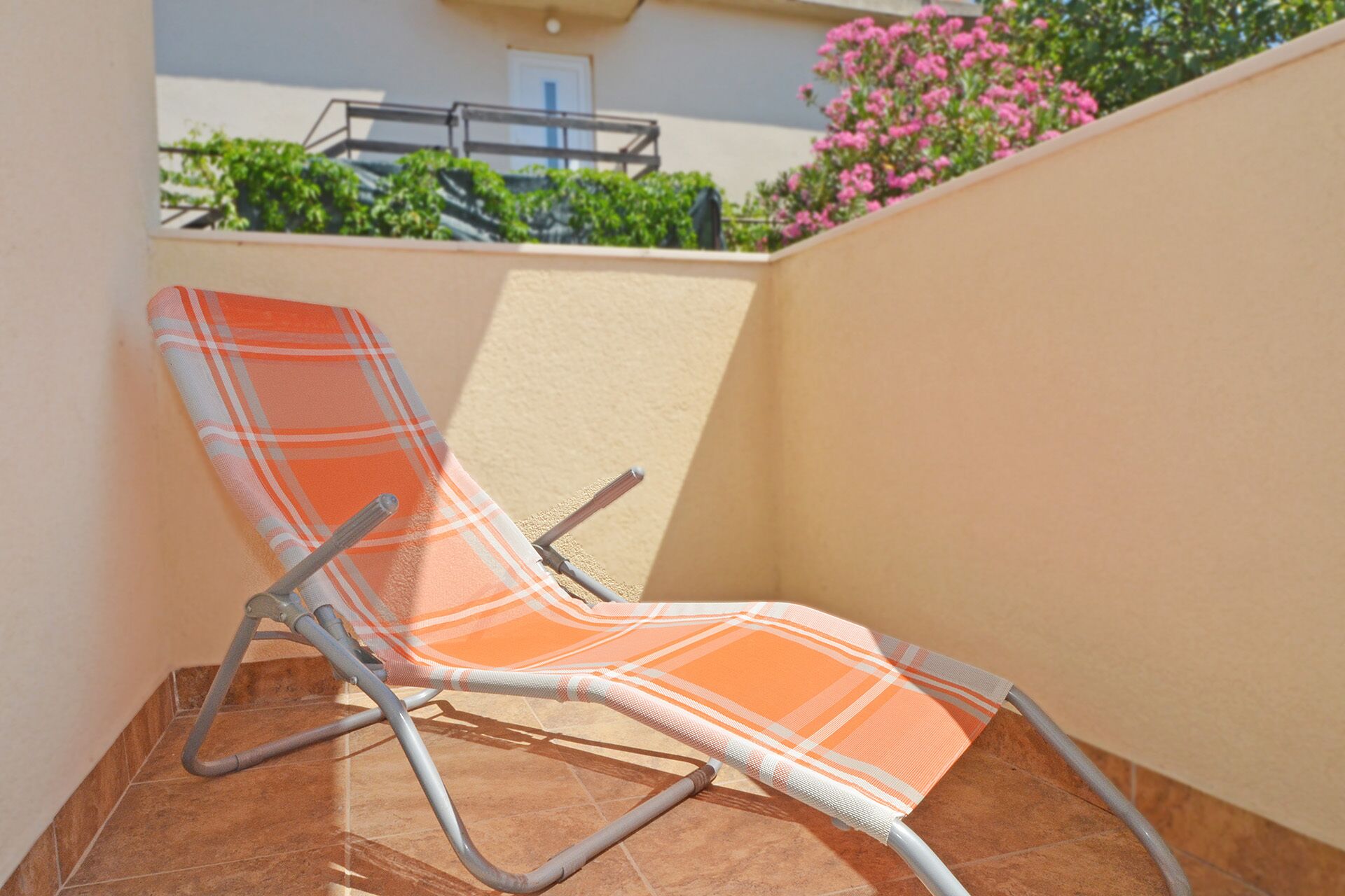 Un seul fauteuil, doté d’un revêtement à carreaux, est placé sur un sol carrelé, à côté d’un mur percé d’une fenêtre donnant sur un jardin.