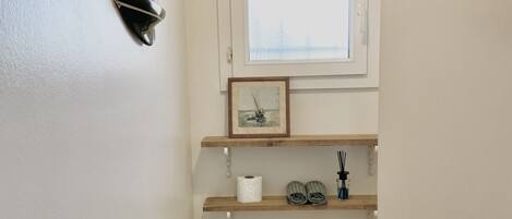 A small, clean bathroom with a toilet, a shelf with toiletries, a framed picture above the window, and a towel rack.