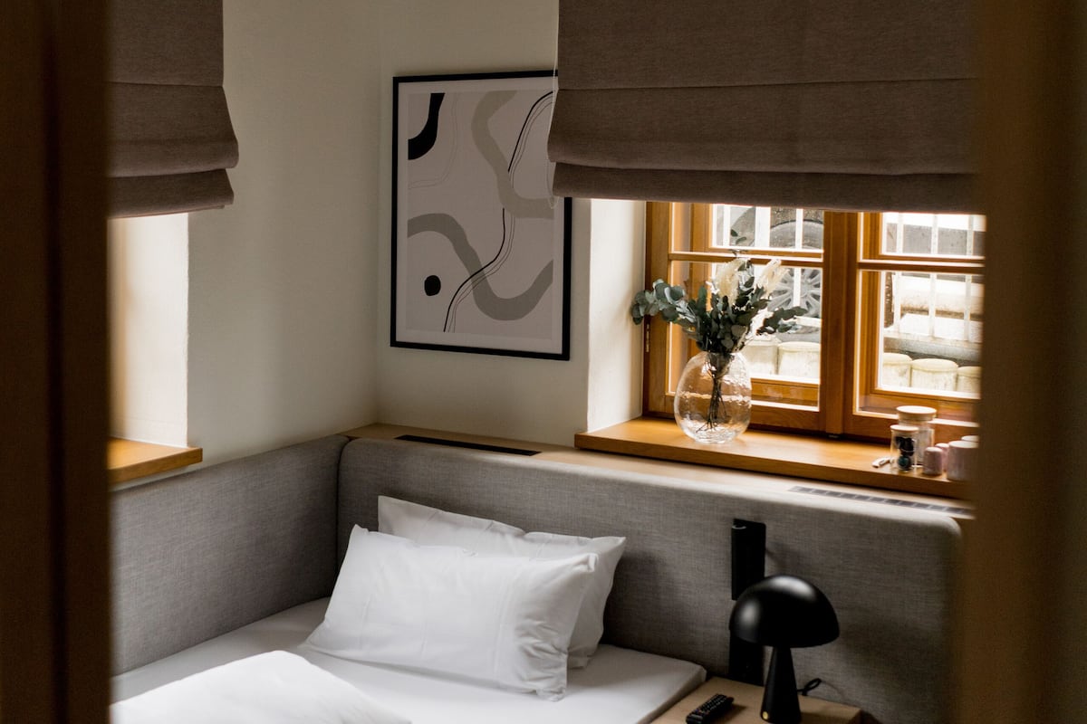 Un lit bien fait, avec du linge de lit blanc, une table de chevet avec une lampe et une fenêtre donnant sur l’extérieur.
