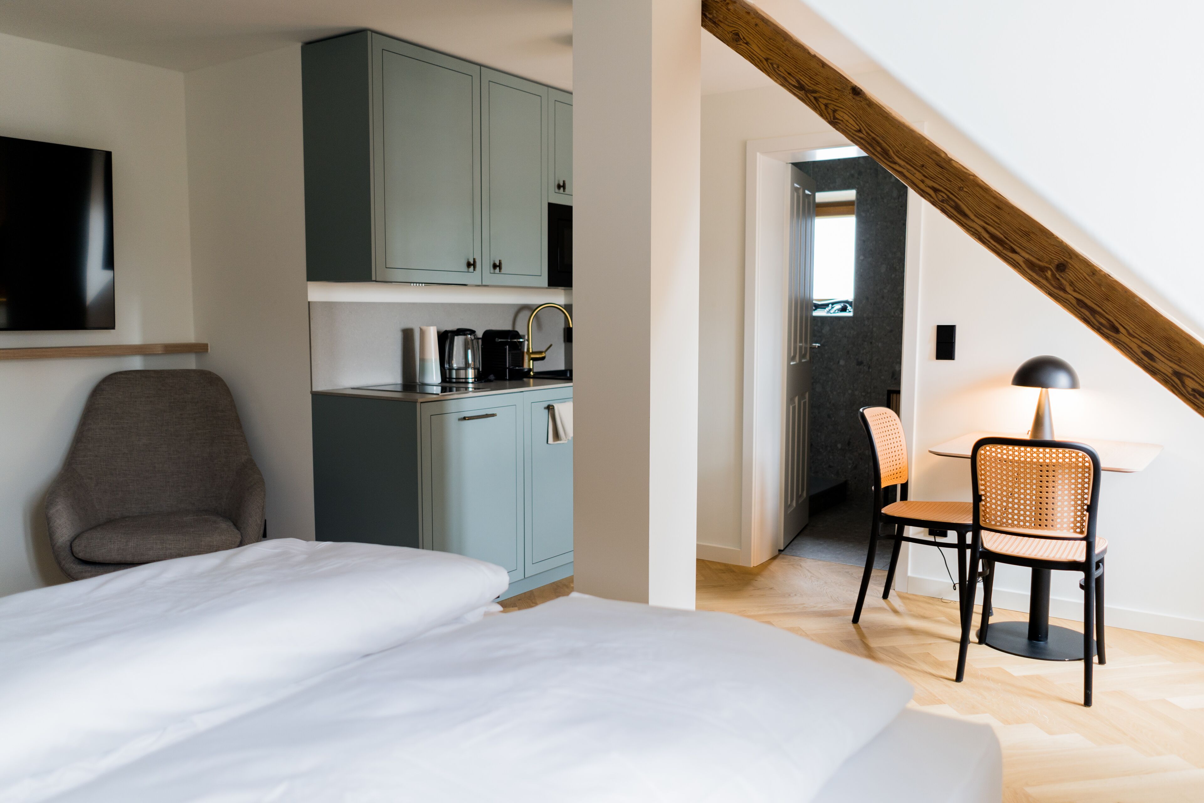 Un dormitorio con cama, una silla, un escritorio con lámpara y una kitchenette con armarios y cafetera.