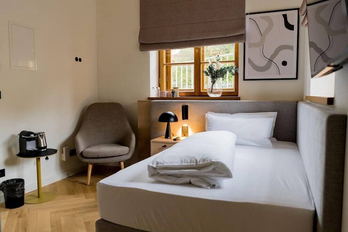 Une chambre d’hôtel avec un lit, une chaise, une table de chevet avec une lampe, une fenêtre donnant sur un paysage et un mur orné d’œuvres d’art abstraites.