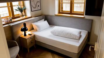 A small, neatly arranged bedroom with a single bed, bedside table, lamp, and a window with curtains.