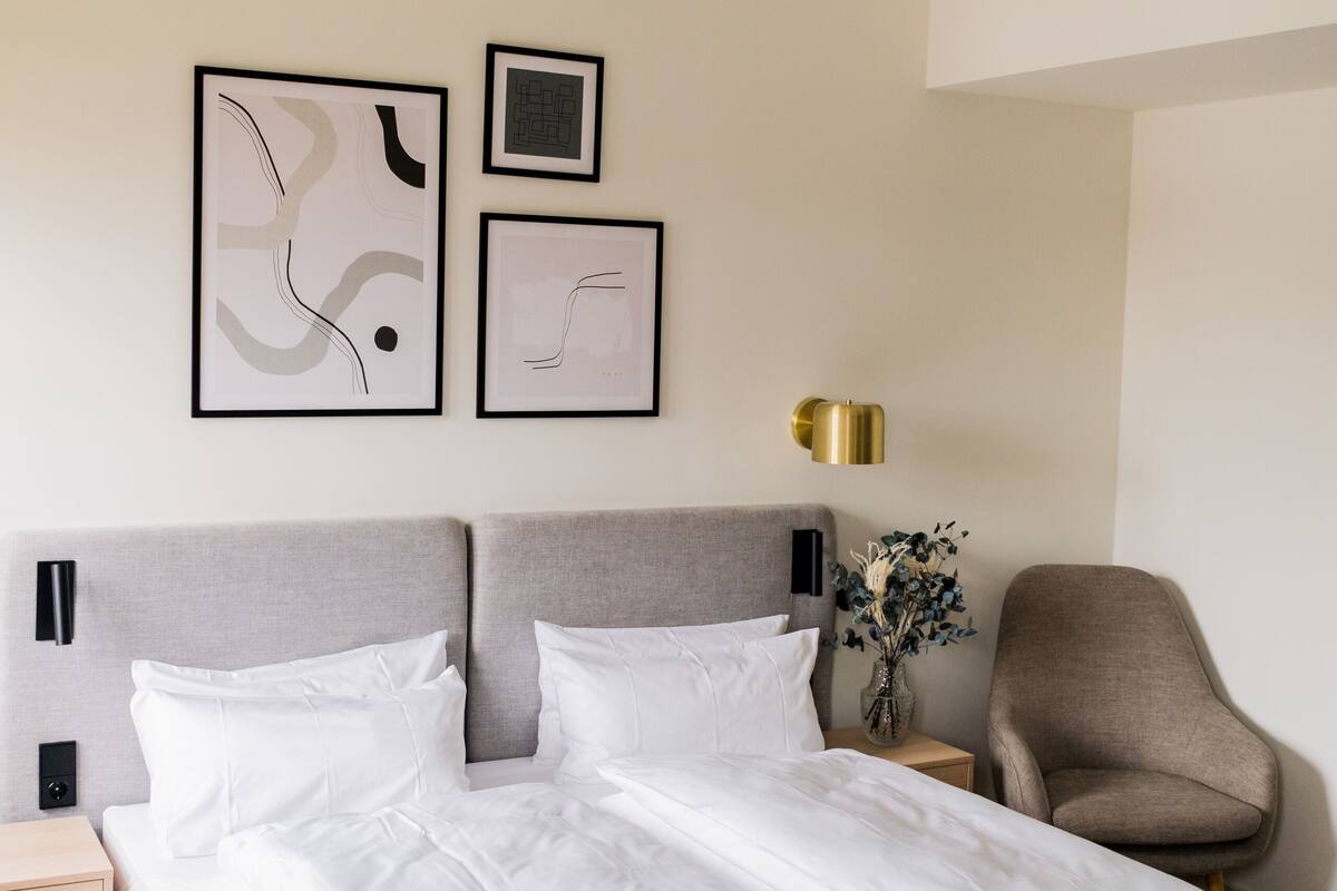 Un lit bien fait, avec du linge de lit blanc, une tête de lit grise et une table de chevet sur laquelle se trouve une lampe. Deux œuvres d’art abstraites encadrées sont accrochées au mur au-dessus du lit.