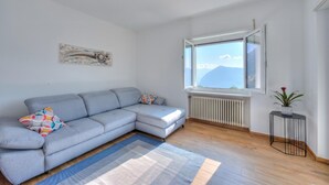 A modern living room with a grey sectional sofa, a blue and white rug, a small side table with a plant, and a window with a mountain view. - Ca' Gina Panoramica - Monte Bre (Lugano)