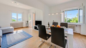 A modern living room with a sofa, a dining table with chairs, a TV, and a large window with a view. - Ca' Gina Panoramica - Monte Bre (Lugano)