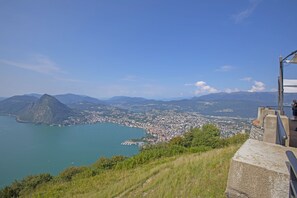 A panoramic view of a coastal city with a prominent mountain, a clear blue sky, and a distant view of a harbor. - Ca' Gina Panoramica - Monte Bre (Lugano)