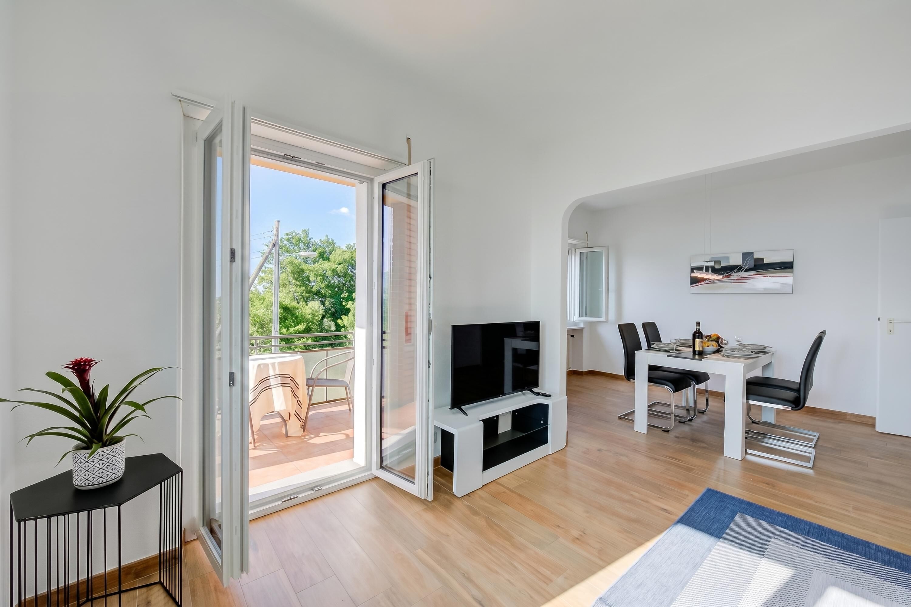 Una sala de estar moderna con televisor, mesa de comedor, sillas y un ventanal corredizo que da a un balcón.