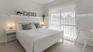 Un dormitorio con una cama, mesitas de noche, una silla, una ventana con cortinas traslúcidas y una planta en la pared.