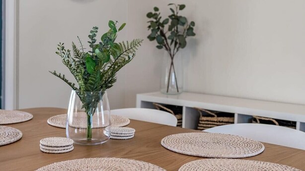 Una mesa de comedor con individuales tejidos, un florero de vidrio con follaje verde y una silla blanca.