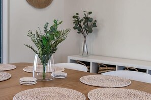 A dining table with woven placemats, a glass vase with green foliage, and a white chair. - Home on Haynes (Goolwa Beach)