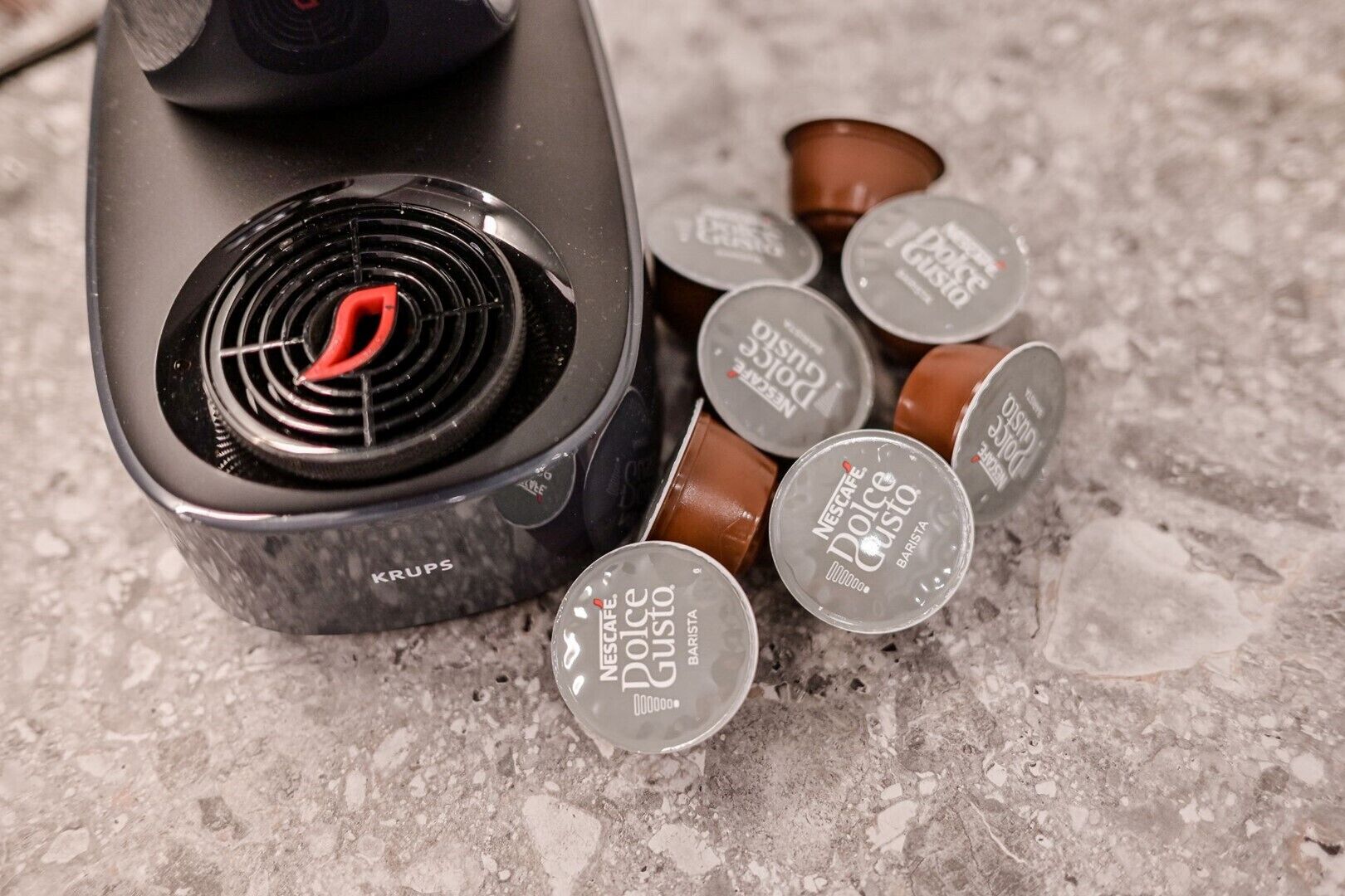A coffee machine with a Krups brand label and Nescafe Dolce Gusto coffee pods on a textured surface.