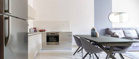 Una cocina moderna con horno empotrado, un comedor con mesa de vidrio y sillas grises, y un sofá gris con un espejo encima.