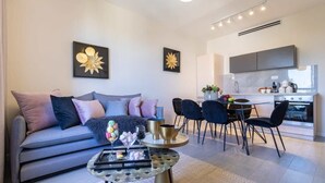 A modern living room with a grey sofa, a coffee table, and a dining area with chairs and a kitchen in the background. - King David Luxury 2BR/Parking/View Old City (Jerusalem)