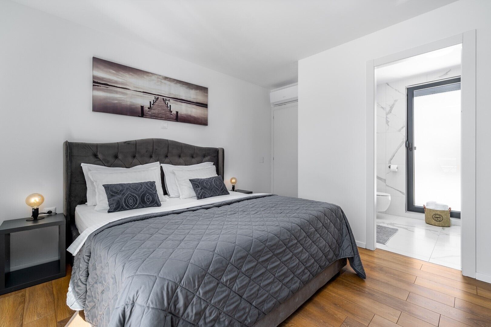 Un dormitorio moderno con una cama grande, una mesita de noche con lámpara, un cuadro en la pared y un baño a la vista a través de una puerta abierta.