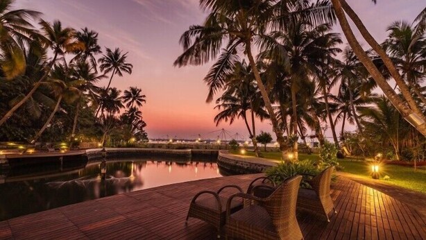 A serene poolside area with a wooden deck, lounge chairs, and palm trees at sunset. - Amã Stays & Trails Sherly'S Ente Kumbalanghi, Kochi (Kochi)