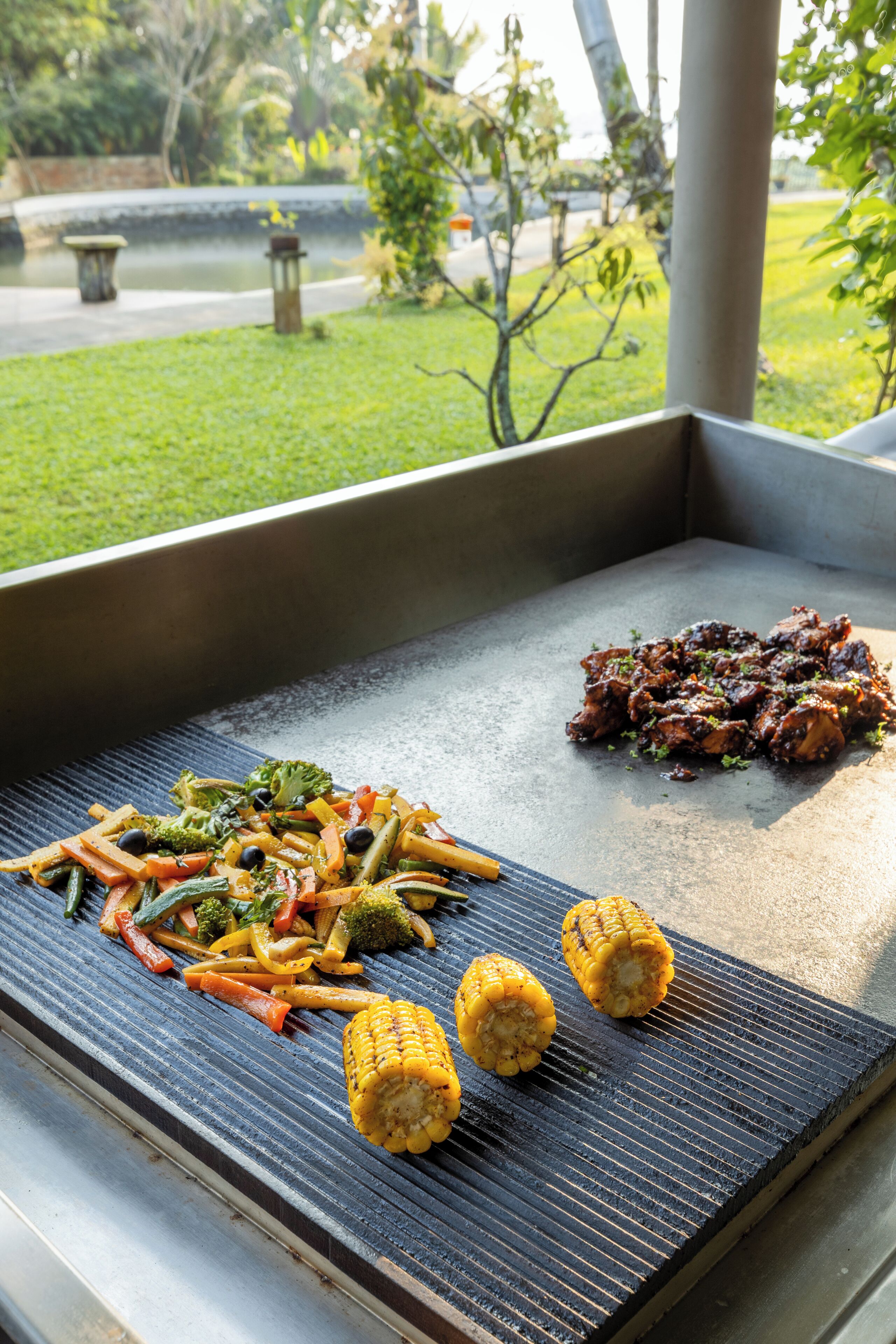 Una griglia con verdure e carne, allestita all'aperto con vista su un giardino.