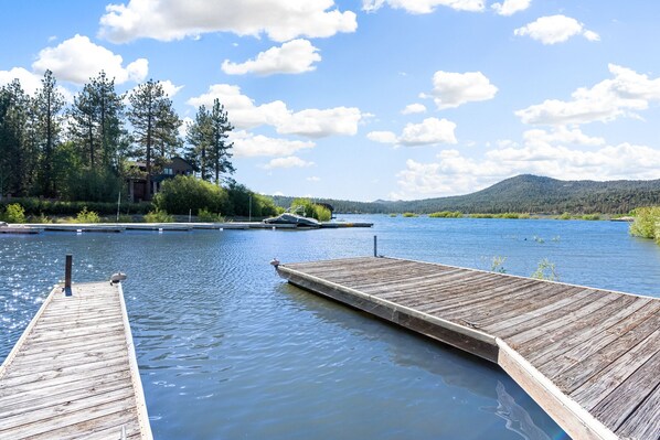 Une jetée en bois s’étendant sur un lac paisible, avec une maison et une forêt en arrière-plan.