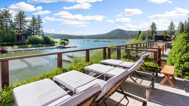 Un patio junto al lago con sillones de descanso y una terraza de madera con vista al lago, rodeado de vegetación y montañas.