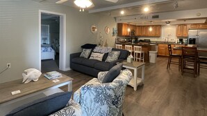 Uma sala de estar moderna com um sofá, mesa de centro e ventoinha no teto. É visível, ao fundo, uma cozinha com armários de madeira e eletrodomésticos de aço inoxidável.