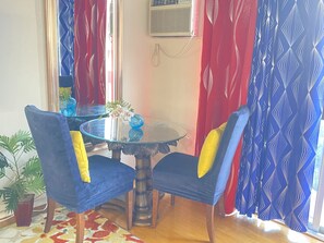 Un comedor con una mesa redonda de cristal, dos sillas azules con cojines amarillos, un florero con flores blancas y una planta en maceta.