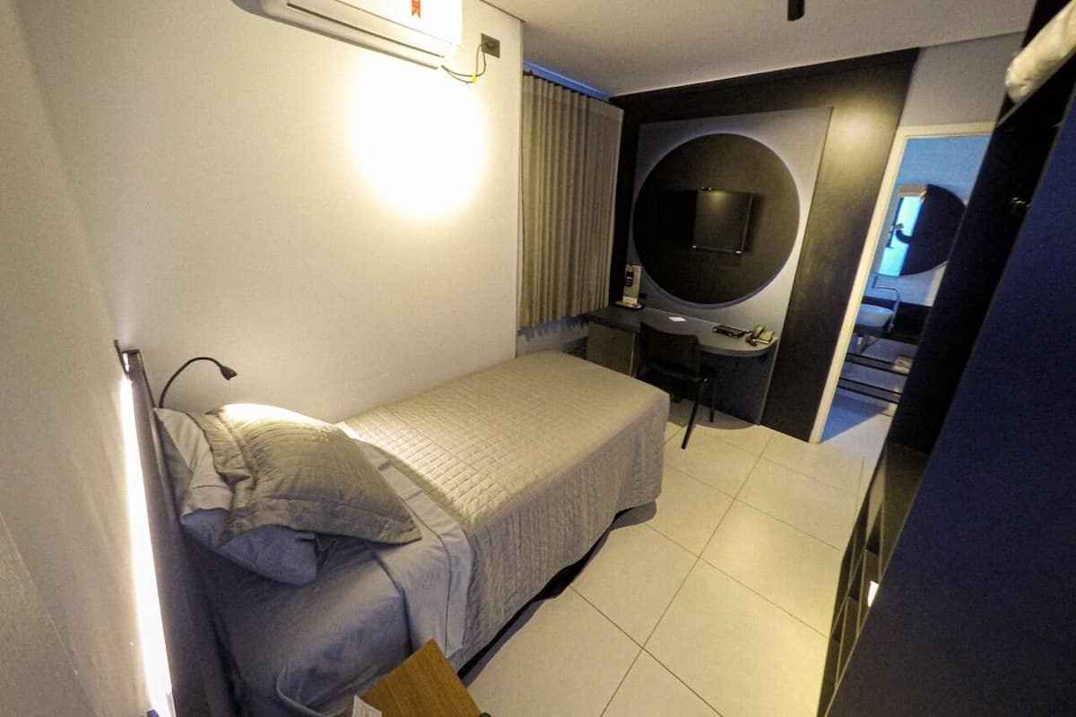 A compact hotel room with a single bed, a desk, a chair, and a round mirror.