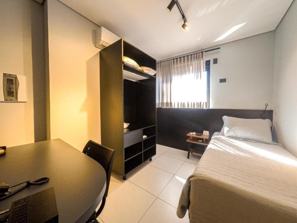 A hotel room with a bed, a desk with a telephone, a bookshelf, and a window with curtains.