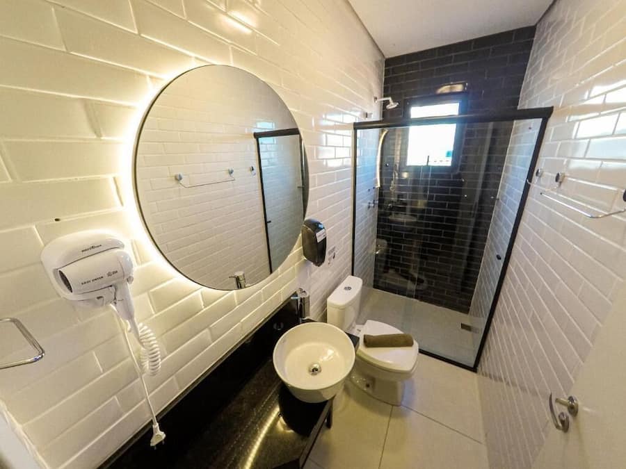 Un baño moderno con pared de ladrillo blanco, un espejo redondo, un lavamanos, un inodoro y una cabina de ducha de vidrio.
