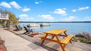 A lakeside picnic area with a wooden table and chairs, a small deck, and boats in the background. - Lakefront Cottage with Hot Tub & Fire Pit (Fife Lake)