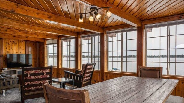 A wooden cabin with a large table and chairs, large windows, and a television. - Lakefront Cottage with Hot Tub & Fire Pit (Fife Lake)