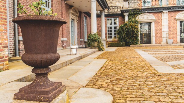 A courtyard with a large planter, cobblestone pathway, and a brick building with columns and balconies. - Domaine Du Vivier L'Agneau (Assesse)