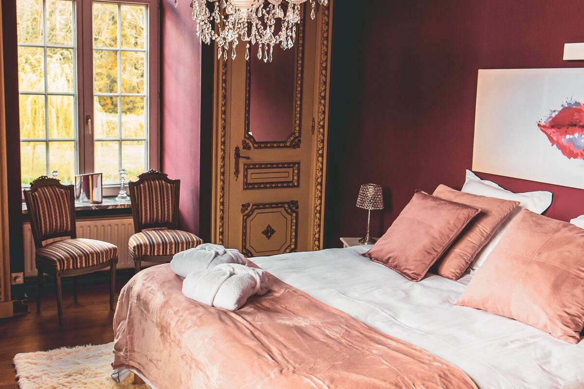 A bedroom with a large bed, a chandelier, a window with a view, and a painting on the wall.
