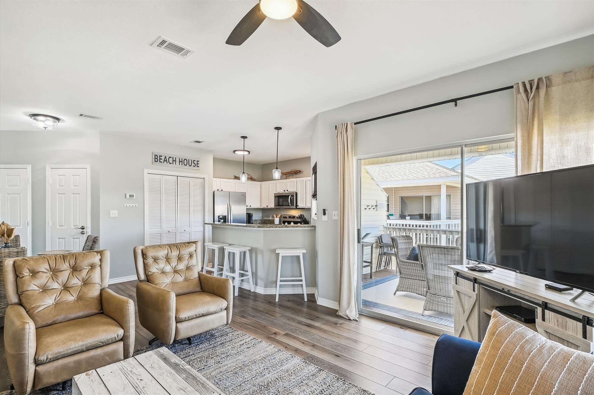 A modern living room with a sofa, armchairs, a coffee table, and a TV. There is a kitchen area with a bar and a dining area with chairs.